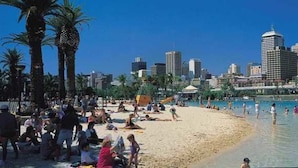 Een strand met een stadsachtergrond, palmbomen en een zwembad.