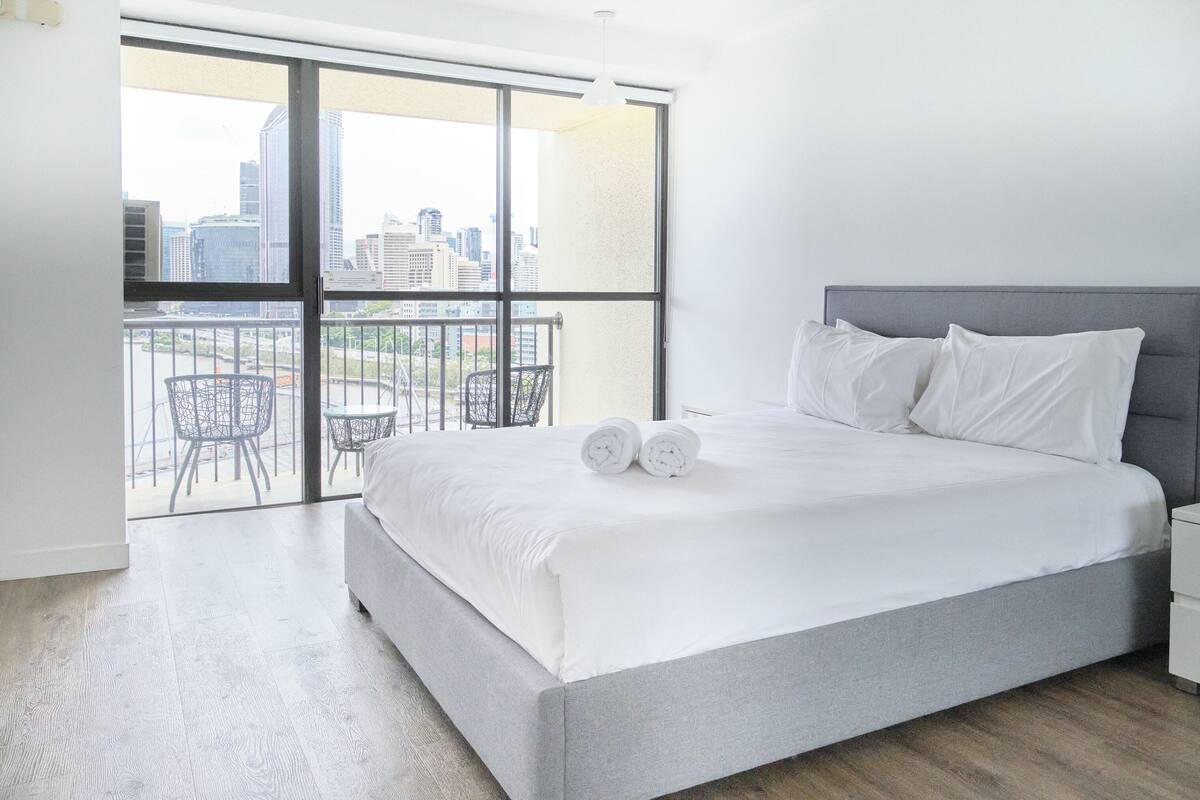 A modern bedroom with a large bed, a grey headboard, and a city view through a balcony.