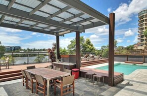 Un espace piscine aménagé sur le toit, avec une table et des chaises en bois, un petit bar et une vue sur une rivière et des immeubles.