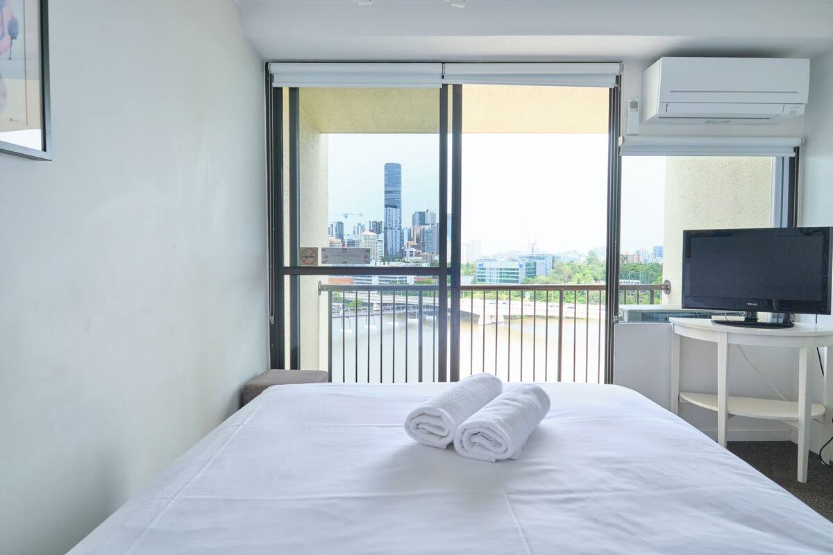 A hotel room with a bed, a TV on a small table, and a city view through the window.