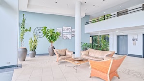 Een moderne lobby met een hoge plafond, een centrale kolom en een tussenverdieping. Er zijn twee banken, een bijzettafeltje en grote potplanten.
