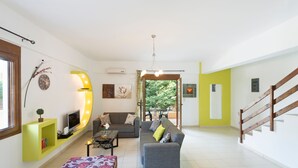 Ein modernes Wohnzimmer mit einem grauen Sofa, einem Couchtisch und einem Fernseher. Eine Treppe befindet sich rechts und ein Fenster links.