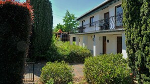 Een gebouw met twee verdiepingen en een balkon, omringd door groen en een stenen muur.