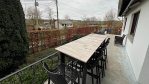 Een balkon met een houten tafel en zwarte stoelen, met uitzicht op een woonwijk met huizen en een haag.