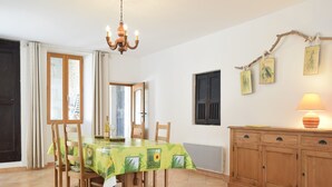 A dining area with a table covered in a sunflower tablecloth, wooden chairs, a chandelier, a cabinet with a lamp, and a window with curtains.