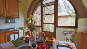 Une table à manger est dressée avec des assiettes, des couverts, un vase rempli de fleurs et une bougie. On peut admirer, à travers une grande fenêtre en arc de cercle, un mur de pierre et la verdure environnante.