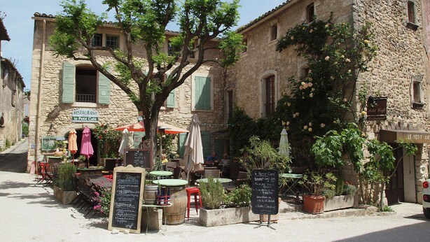 A rustic stone building with a café named "Le Petit Café" featuring outdoor seating and greenery.