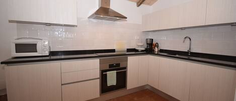 A kitchen with light wood cabinets, a black countertop, a microwave, an oven, a sink, and a stainless steel range hood.
