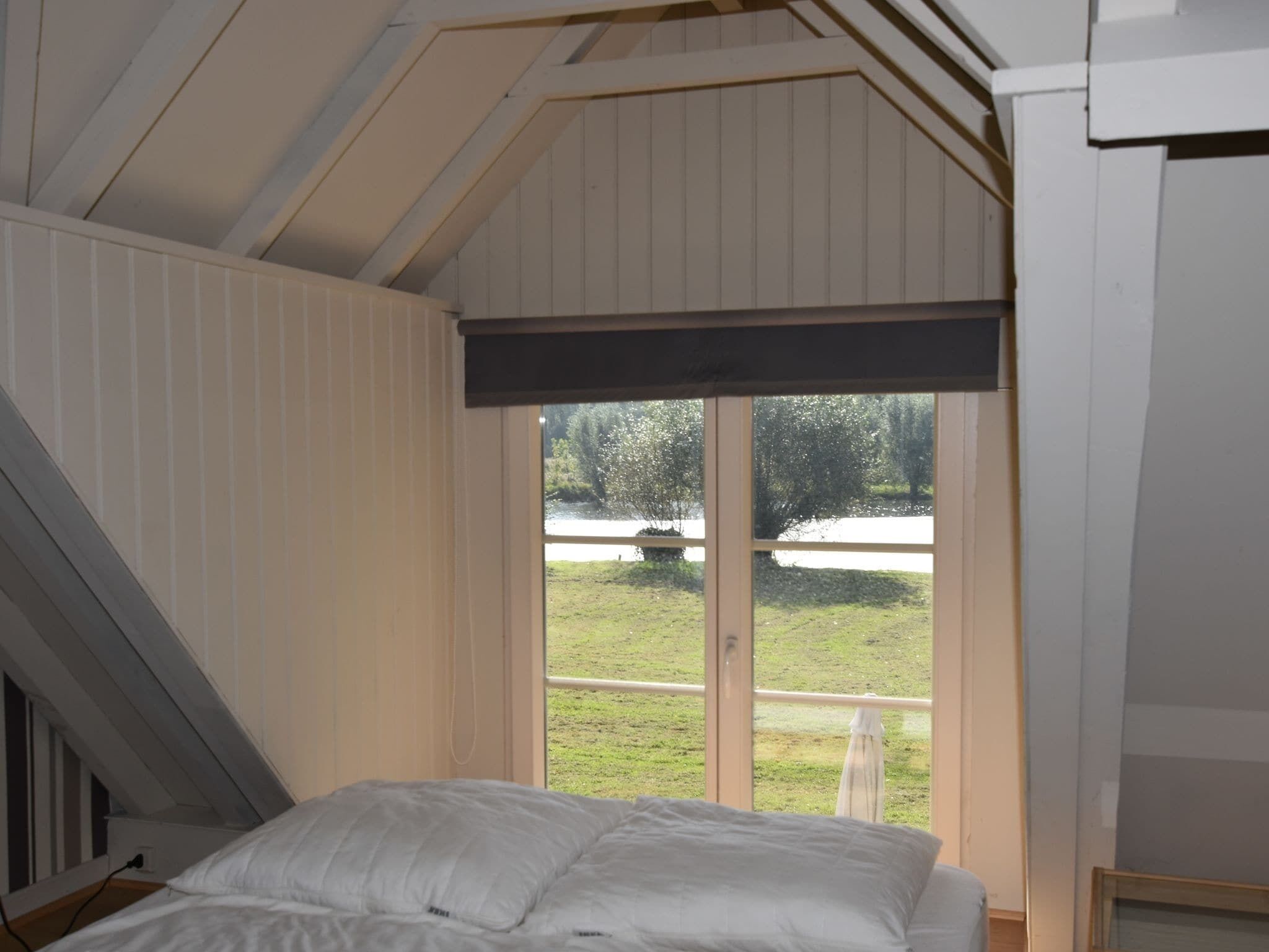 A bedroom with a sloped ceiling, a large window, and a view of a grassy area and trees.