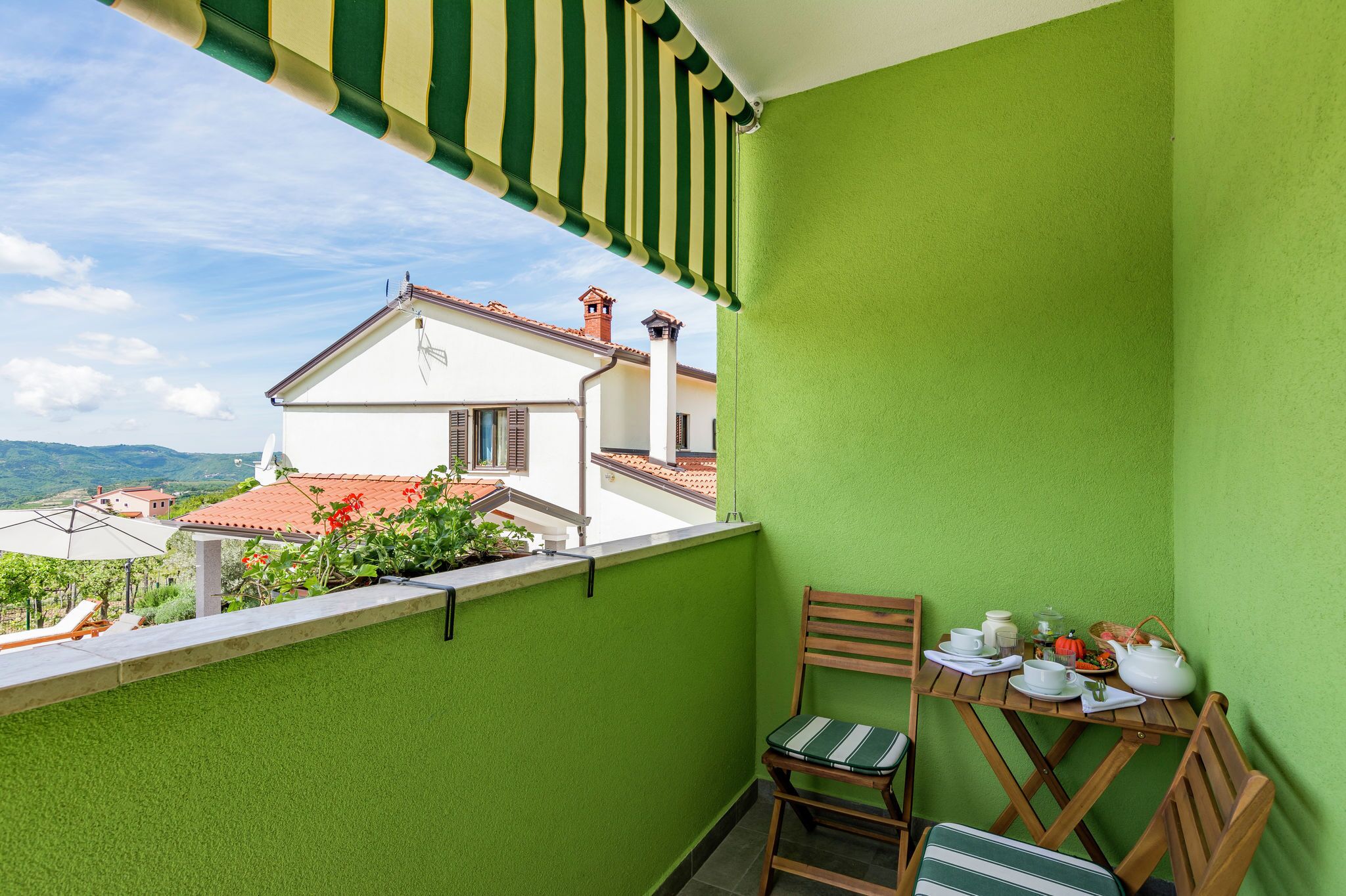 A balcony with a green wall, a wooden table with a chair, and a view of a white building with a red roof.