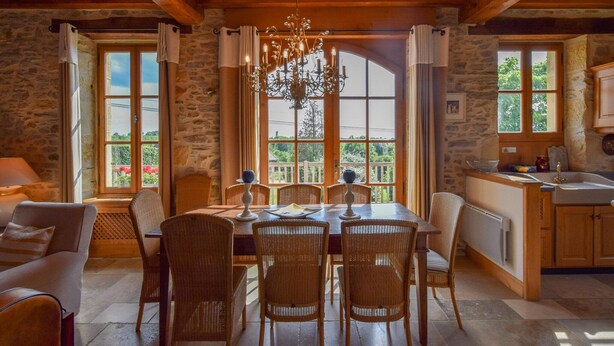A rustic dining area with a stone wall, wooden beams, wicker chairs, a chandelier, and a view of greenery through large windows. - Elegant Villa in Saint-Médard (St. Medard D'excideuil)