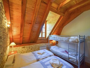Une chambre confortable située dans les combles, avec des poutres en bois, un mur en pierre et une fenêtre. On y trouve deux lits et une lampe.