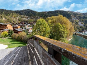 Une terrasse en bois donnant sur une rivière, avec des bâtiments traditionnels en bois et une colline boisée en arrière-plan.