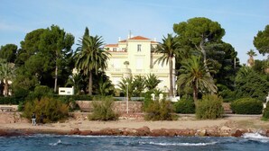 A coastal building with a balcony, surrounded by palm trees and a rocky shoreline.