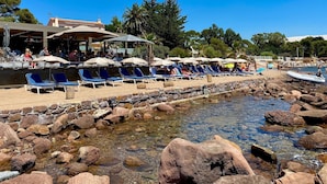 A beachside resort with lounge chairs and umbrellas, a rocky shoreline, and a clear blue sky.