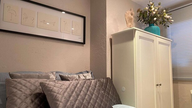 Un dormitorio con cama, almohadas, un cuadro enmarco, un armario y un jarrón con flores.