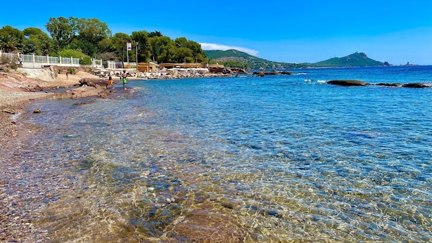 A beach with clear shallow waters, rocky shore, and a distant hill.