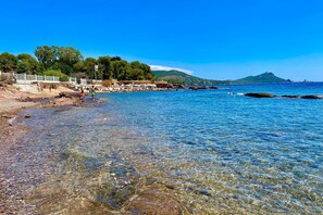 A coastal beach scene with clear water, a rocky shoreline, and a distant mountain.