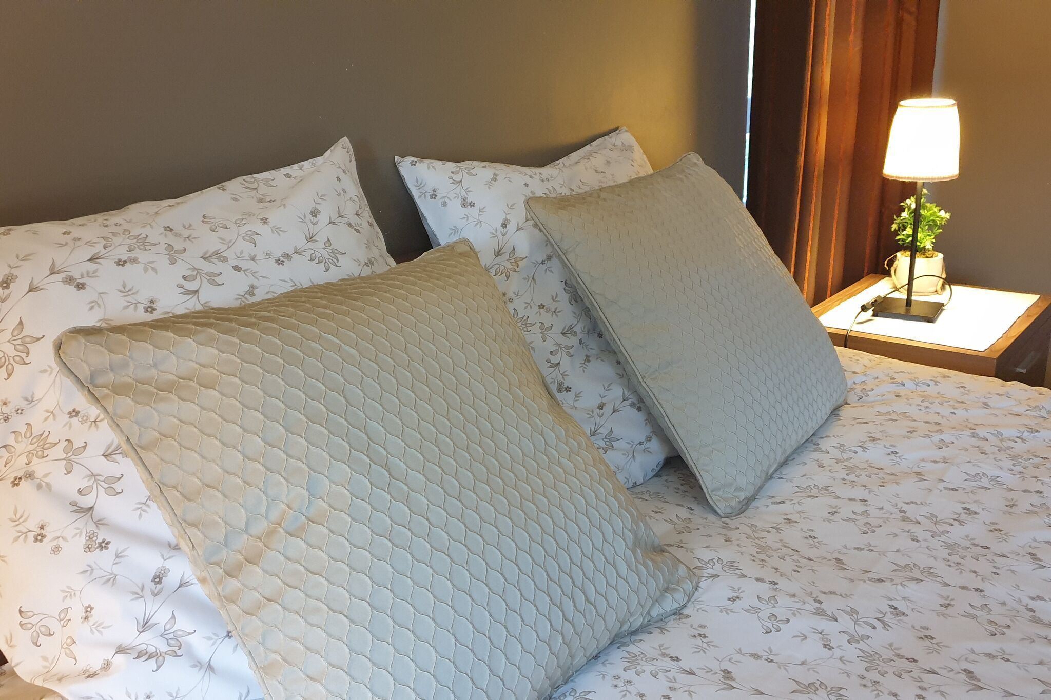 Uma cama bem arrumada com roupa de cama floral e duas almofadas, um abajur e uma pequena planta em vaso sobre uma mesa de cabeceira de madeira.