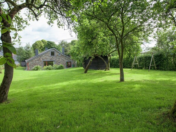 A stone house with a sloped roof, a wooden barn, and a swing set in a grassy yard.