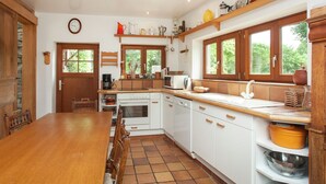 Een keuken met houten kastjes, een betegelde vloer, een houten eettafel en een raam met uitzicht op groen.