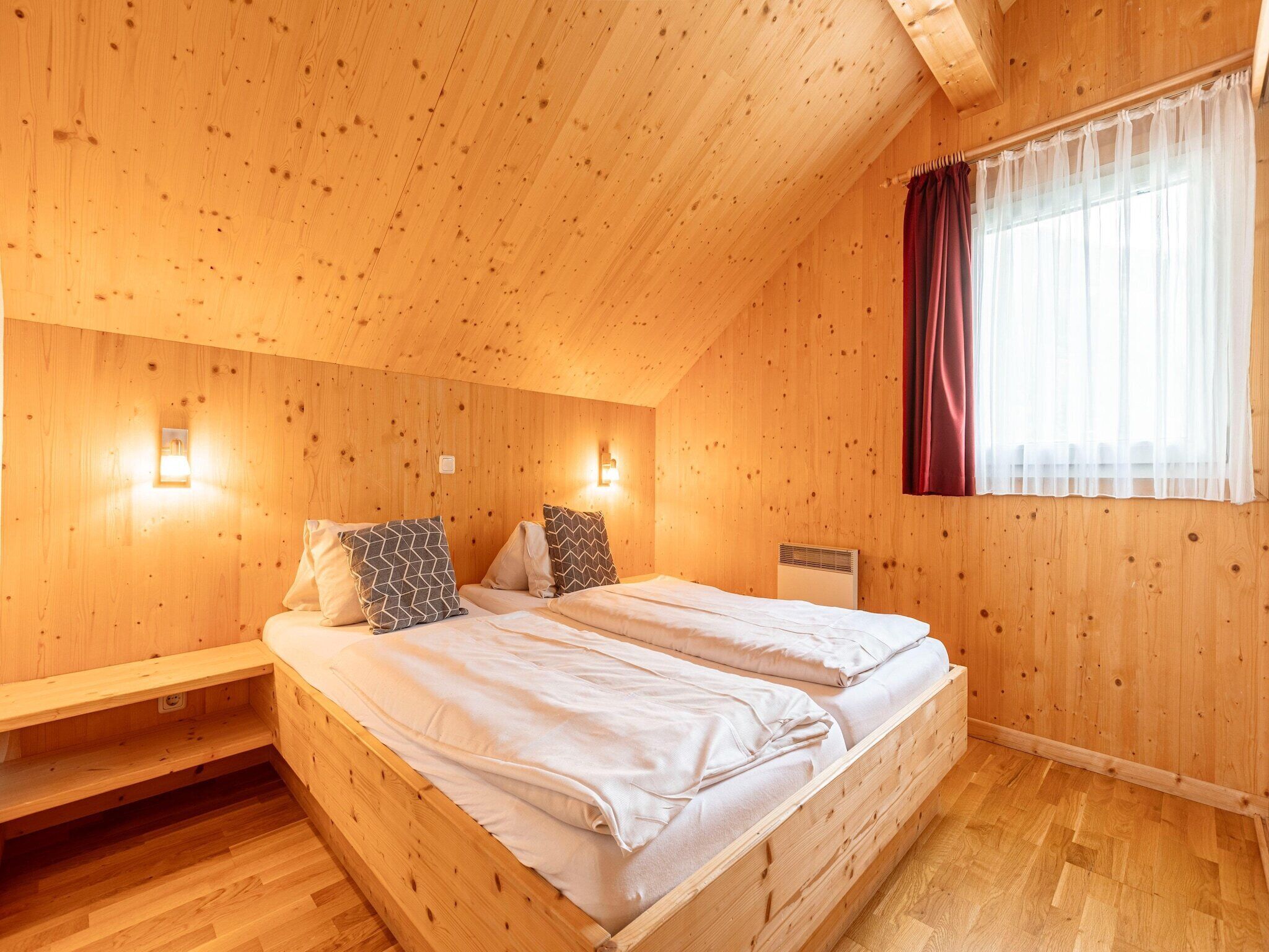 A wooden room with a bed, two bedside tables, a window with curtains, and wall-mounted lights.