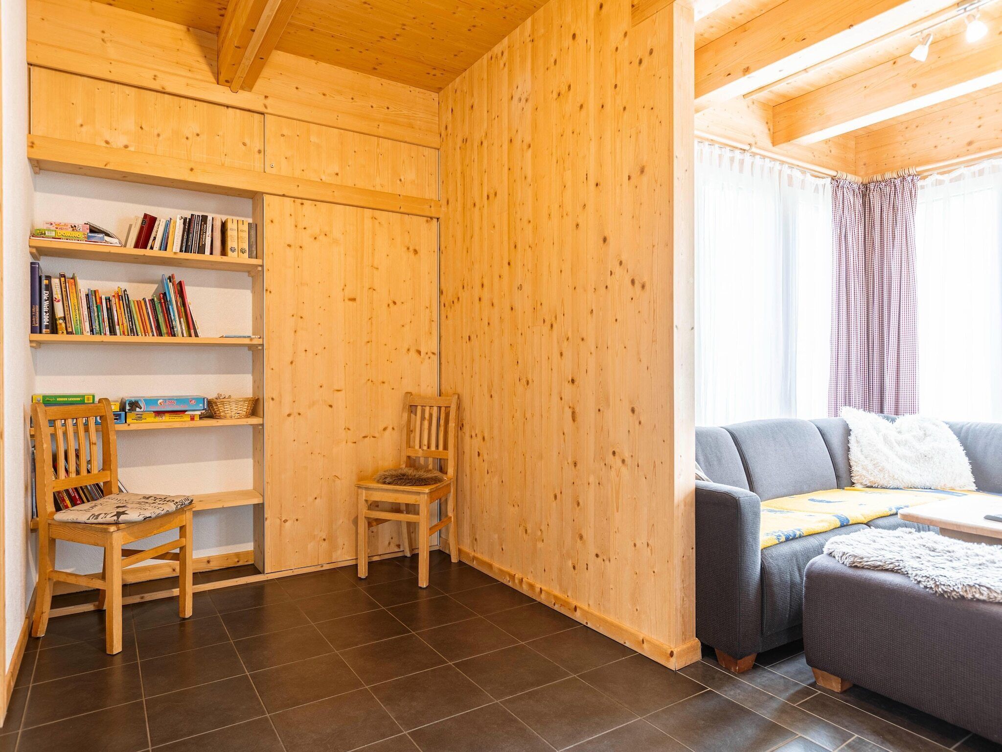 Una habitación acogedora con paredes de madera, una estantería, dos sillas, un sofá y una ventana con cortinas.
