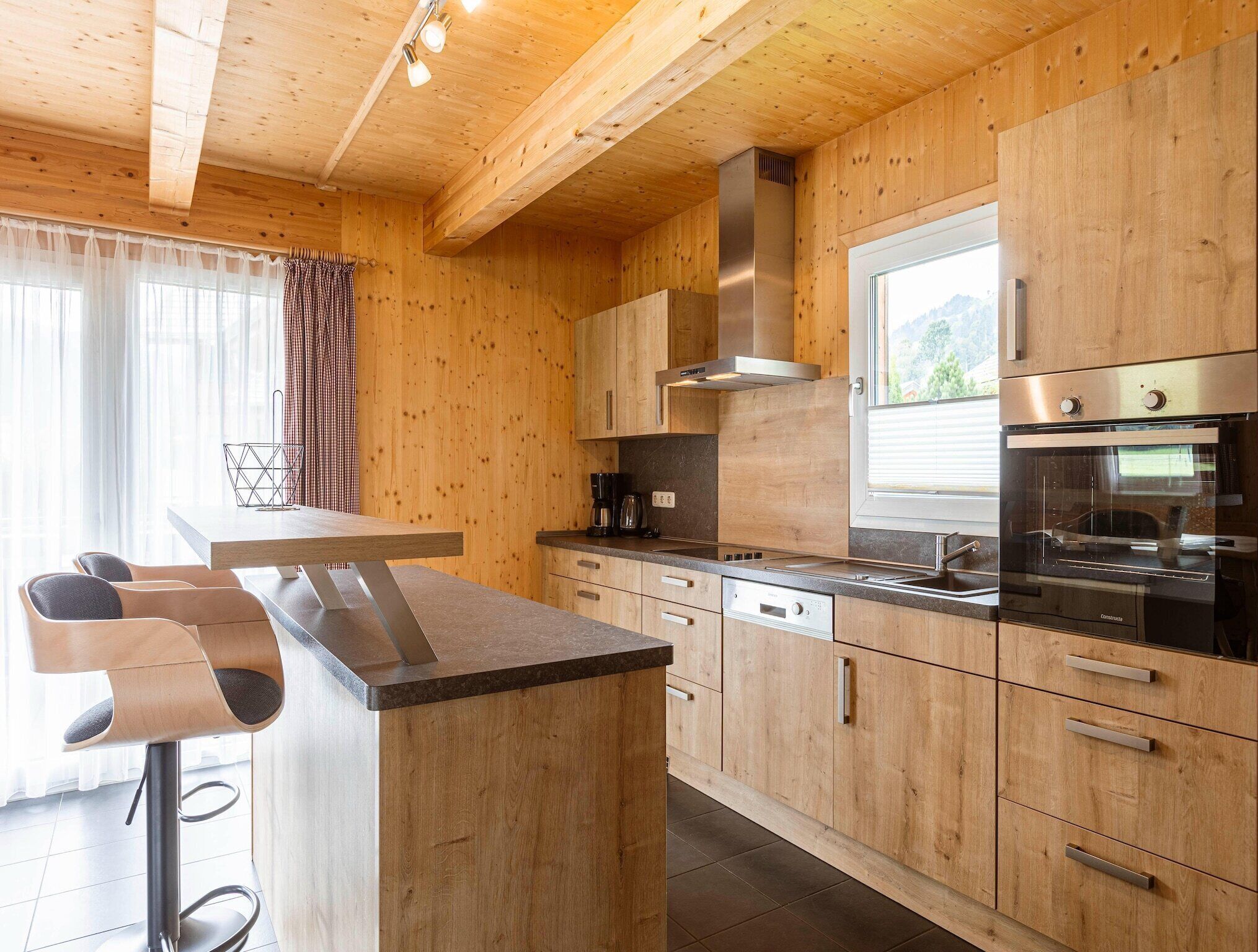 Une cuisine moderne avec des meubles en bois, une hotte en acier inoxydable, un four encastré et un coin bar avec des tabourets.