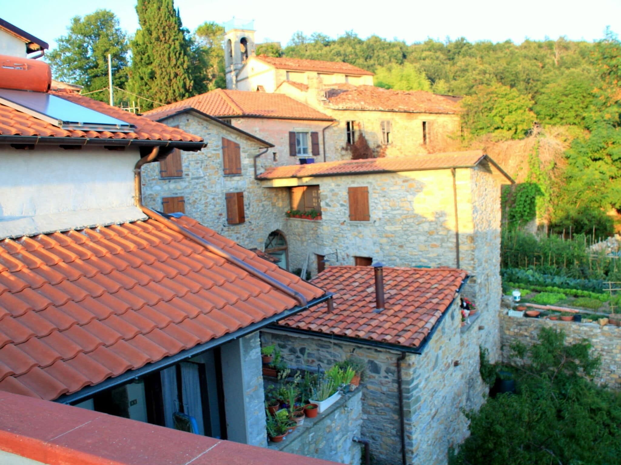 A stone building with a red-tiled roof, multiple windows, and a garden area with potted plants.