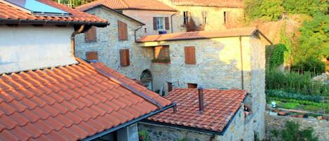 A stone building with a red-tiled roof, multiple windows, and a garden area with potted plants.