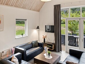Un moderno salón con un sofá de cuero, una mesa de centro, un televisor y vistas a un jardín a través de amplias ventanas.