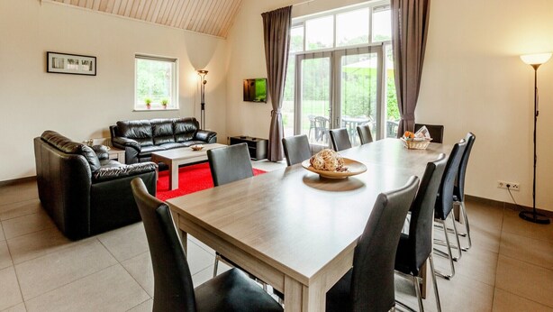A modern living room with a sofa, a coffee table, and a dining table with chairs. There is a TV mounted on the wall, a window with curtains, and a ceiling with wooden beams. - Kempen Comfort with Sauna & Tub (Vessem)