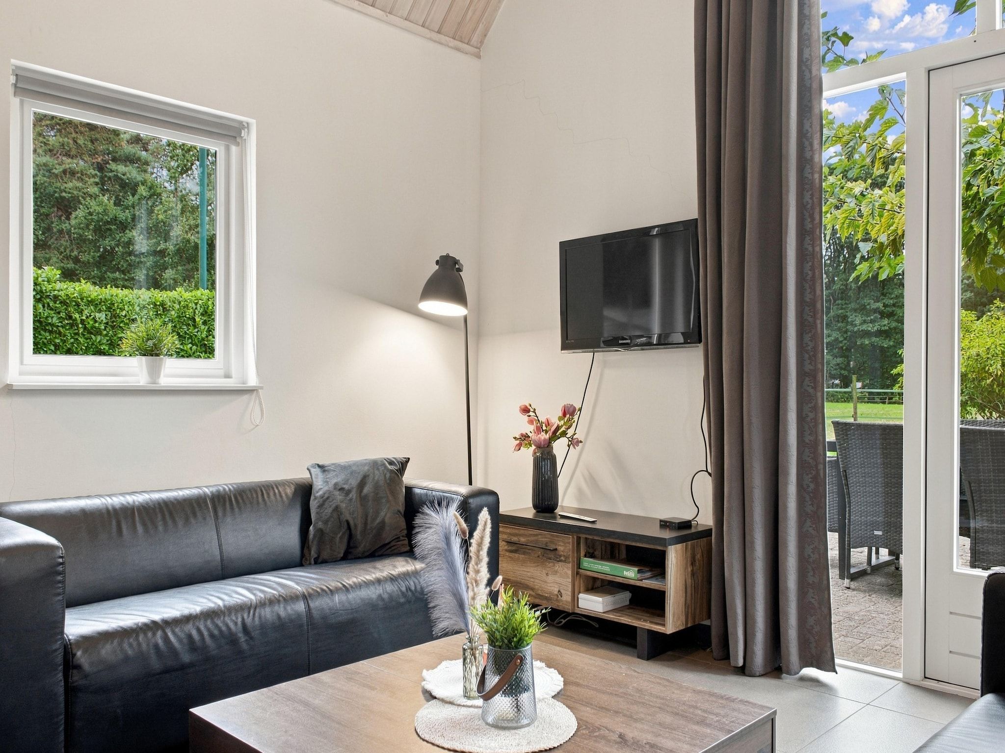 A modern living room with a leather sofa, a coffee table, a TV mounted on the wall, and a view of the outdoors through a sliding glass door.