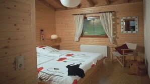 Dormitori d'una cabana de fusta amb llit, una cadira, una taula petita, una finestra amb cortines i una làmpada penjant.