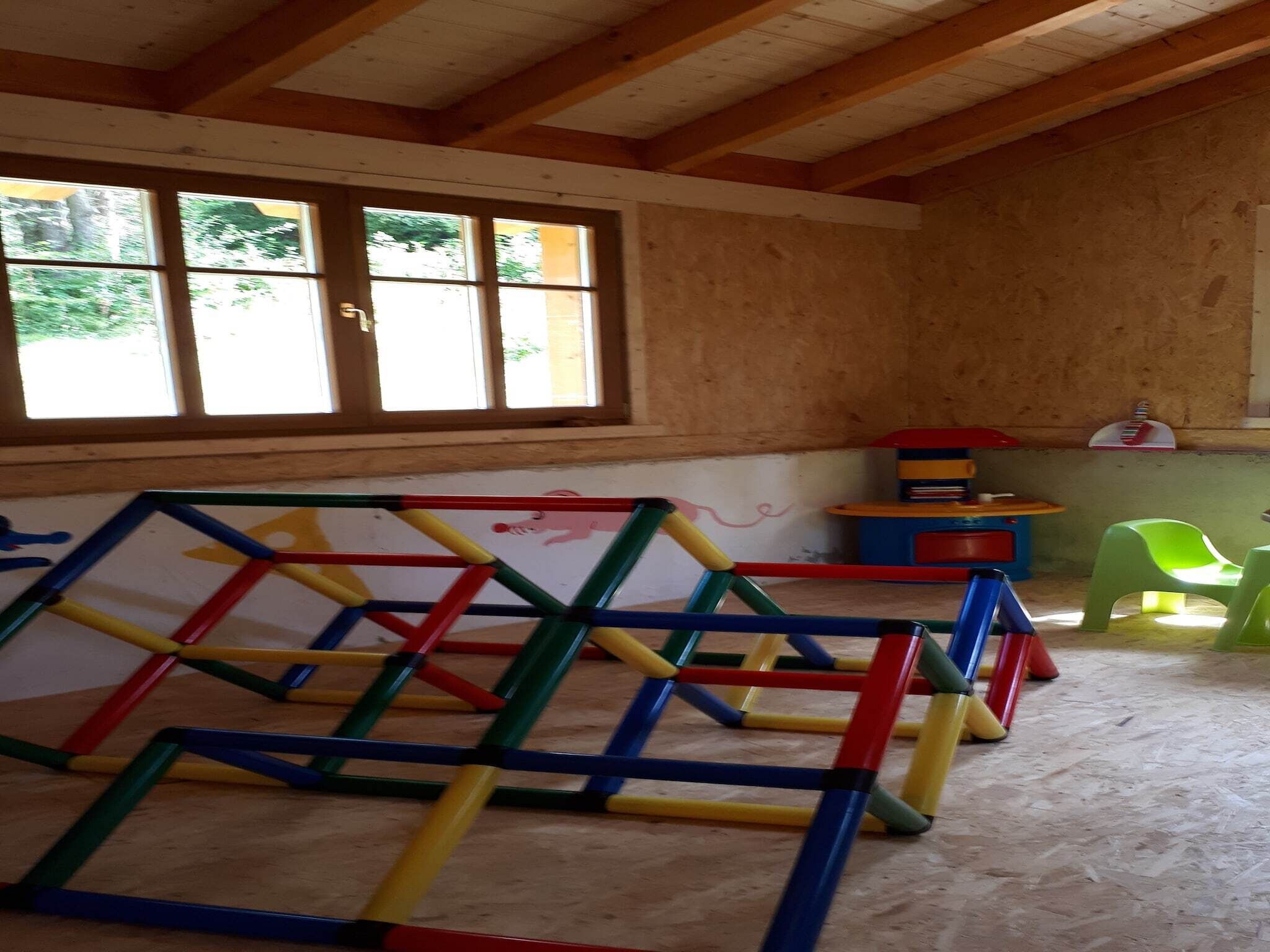 A room with a wooden ceiling, a window with blinds, a colorful play structure, and a small table with chairs.