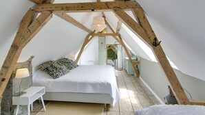 A bedroom with a sloped ceiling, wooden beams, a bed with a white bedspread, a bedside lamp, and a hanging light fixture. - Picturesque fisherman's house near the beach (Scheveningen)
