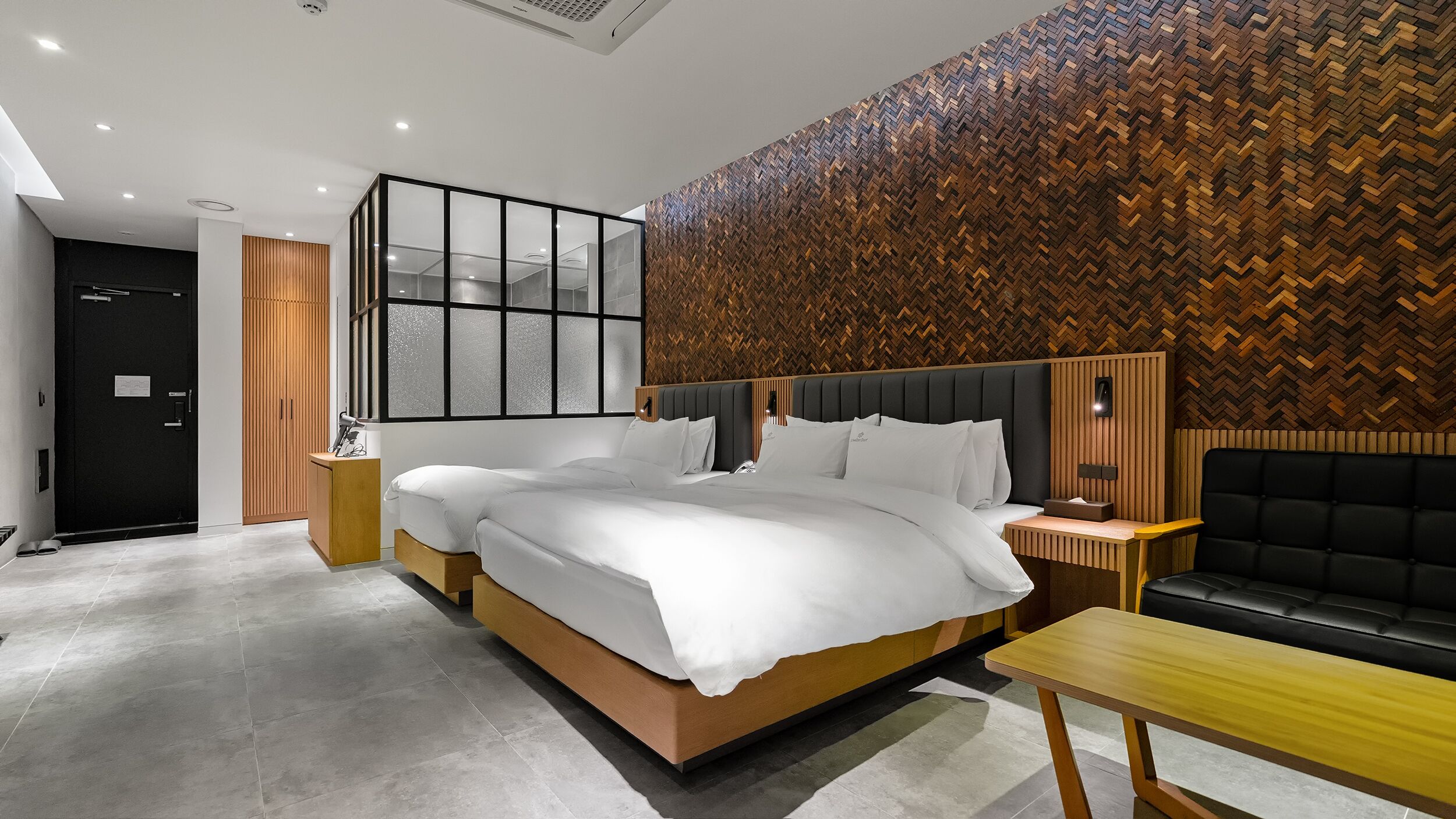 A modern hotel room with a large bed, a wooden headboard, a wooden wall panel, a small table, and a black sofa.