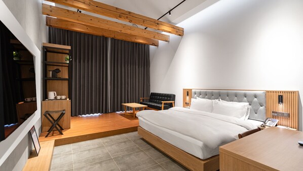 A modern bedroom with a bed, a wooden nightstand, a small table, and a sofa. There is a large window with curtains and a wooden ceiling beam. - Schweizer Dorf Hotel (Cheonan)