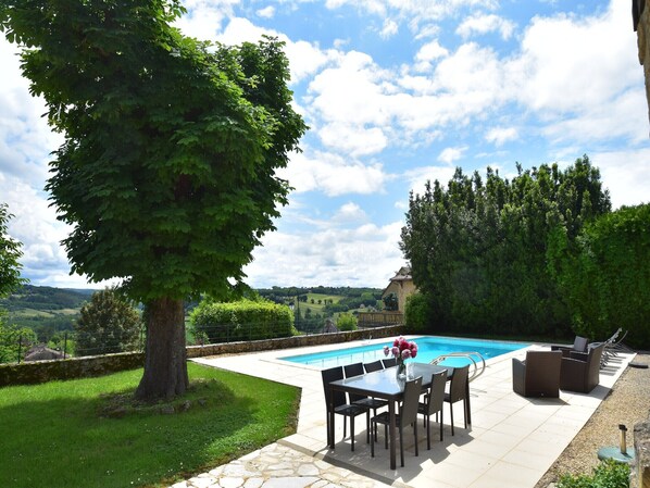 A poolside area with a table and chairs, surrounded by a stone patio and a view of a swimming pool.