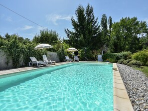 A clear swimming pool with lounge chairs and umbrellas, surrounded by greenery and a white picket fence.