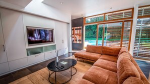 A modern living room with a large flat-screen TV, a sectional sofa, a coffee table, and a view of the outdoors through large windows.