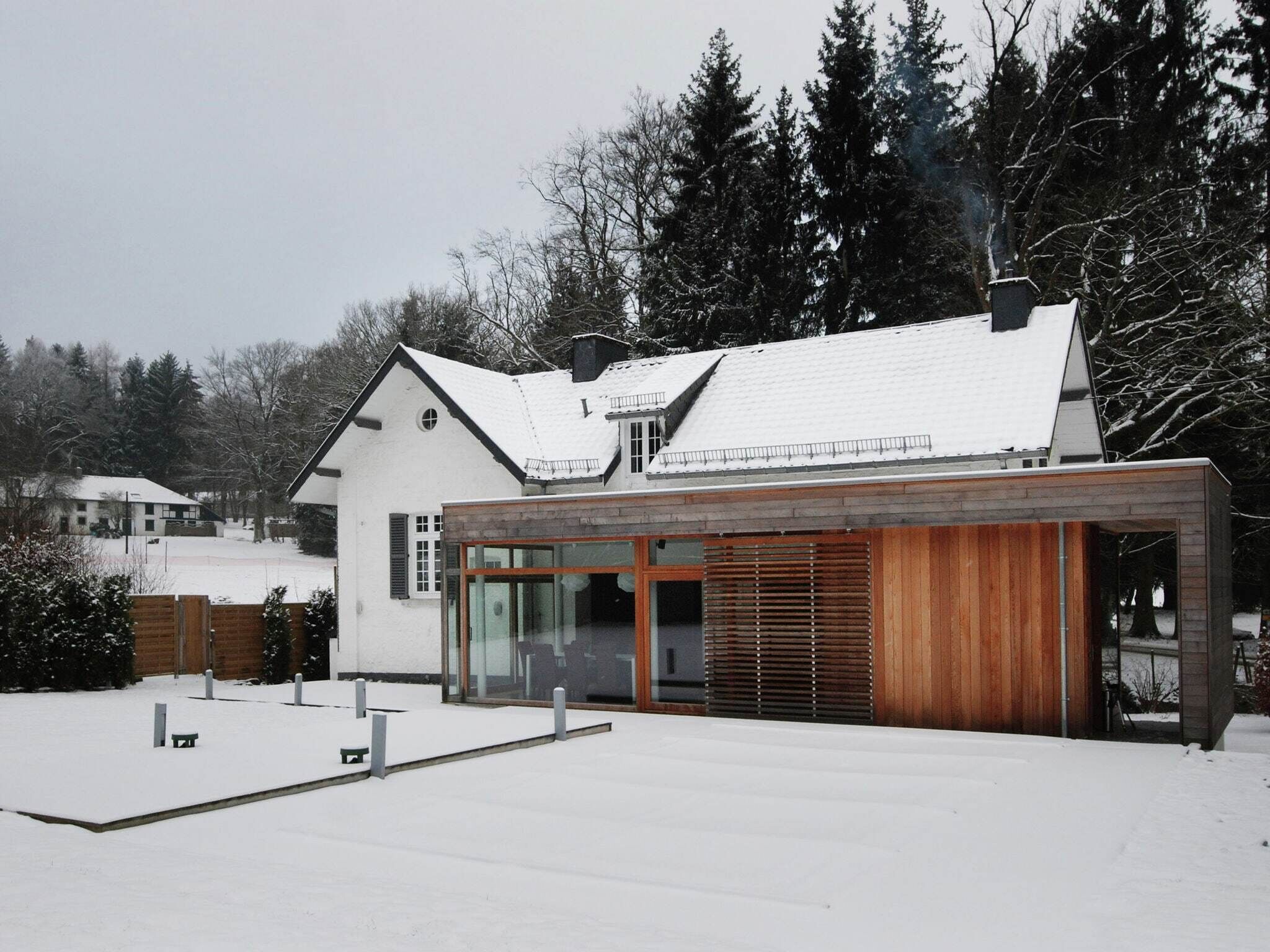 A modern house with a large glass extension and a wooden facade, surrounded by snow-covered ground and trees.