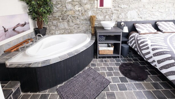 A modern bathroom with a stone wall, a bathtub, a sink, and a bed with patterned bedding.
