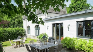 A patio with a wooden table and chairs, surrounded by greenery and a white building with arched windows.