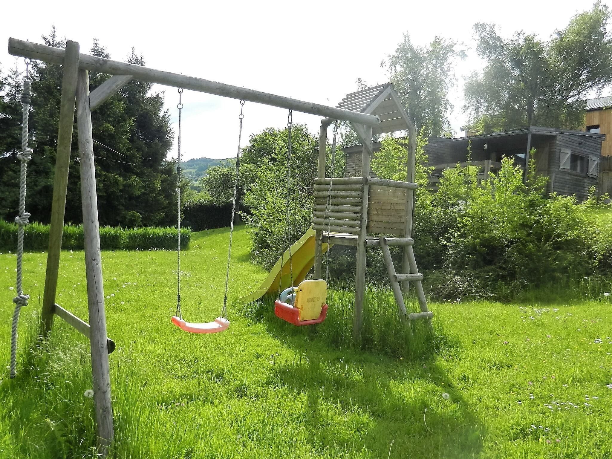 A wooden swing set with a slide, surrounded by green grass and trees.