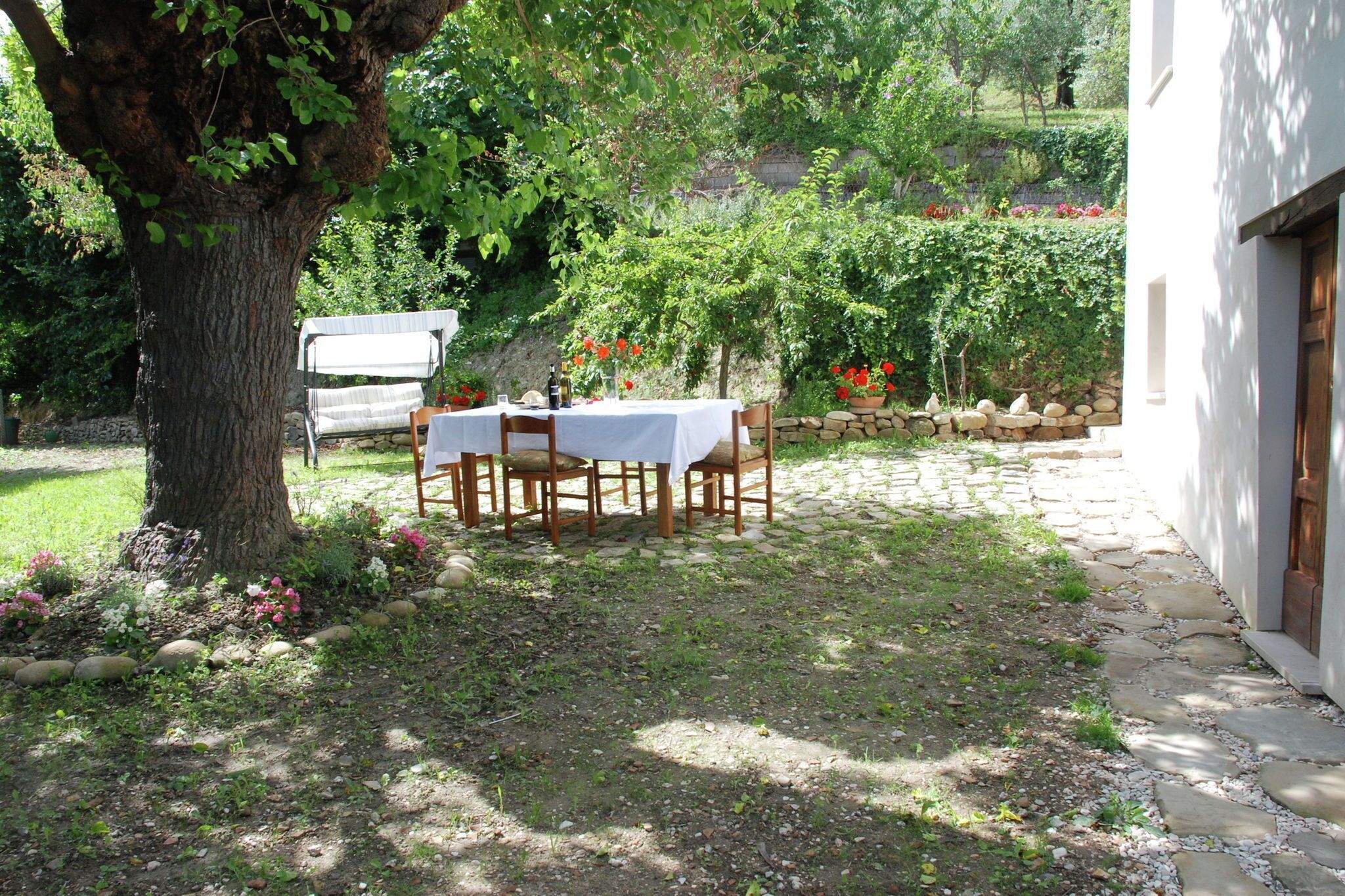 A stone-paved patio with a table set for two, surrounded by lush greenery and a large tree.