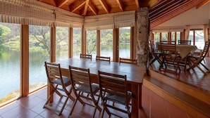 A dining area with a large table and chairs, large windows with a view of a lake, and a wooden ceiling.