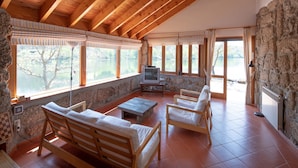 A spacious room with a sloped wooden ceiling, stone walls, and large windows offering a view of a lake. There are wooden furniture pieces, including a sofa and chairs, and a television.