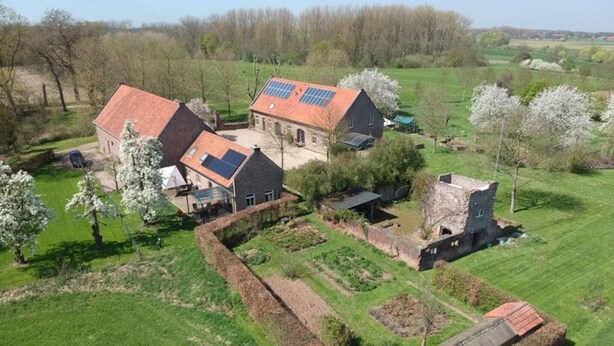 Une propriété rurale comprenant plusieurs bâtiments, dont une maison avec un toit rouge et des panneaux solaires, entourée de champs verdoyants et d’arbres.
