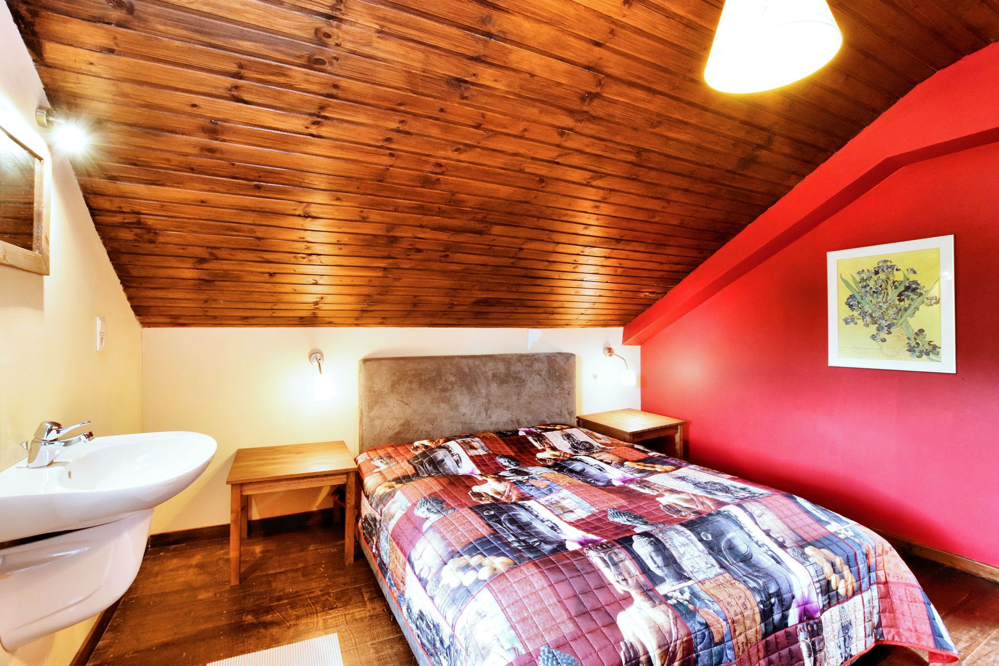 Un dormitorio con techo de madera, pared de acento roja, cama con colcha colorida, mesita de noche y lámpara de pared.